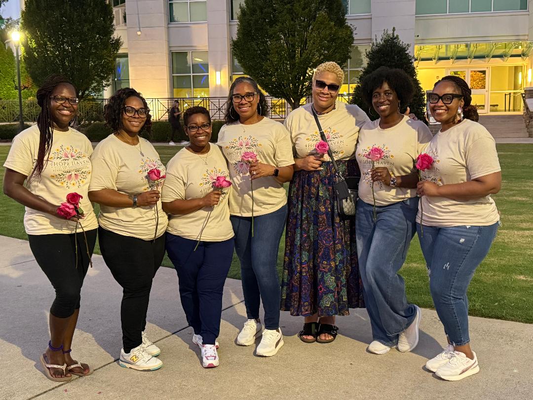 Seven women standing outdoors holding pink flowers, smiling, in front of trees and a building, daytime.