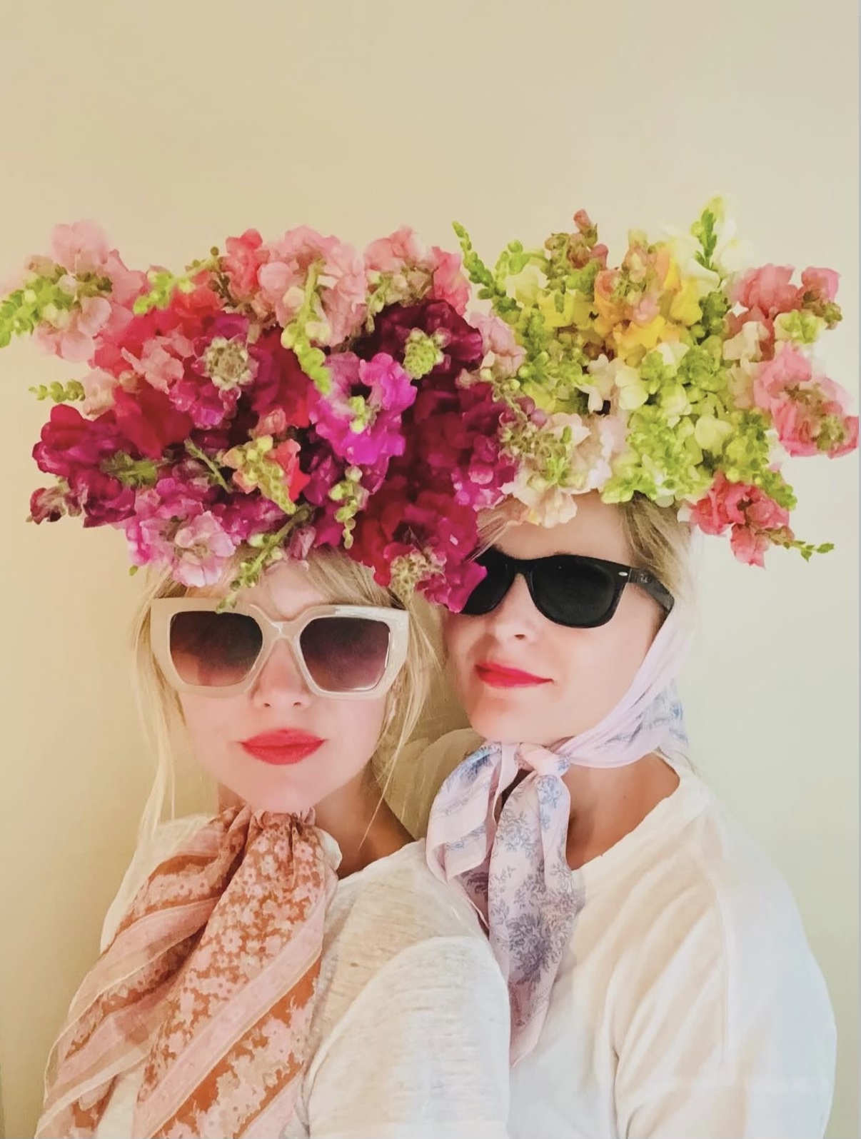 Two women wearing large floral headpieces, sunglasses, and light-colored clothing, standing close together against a plain background.