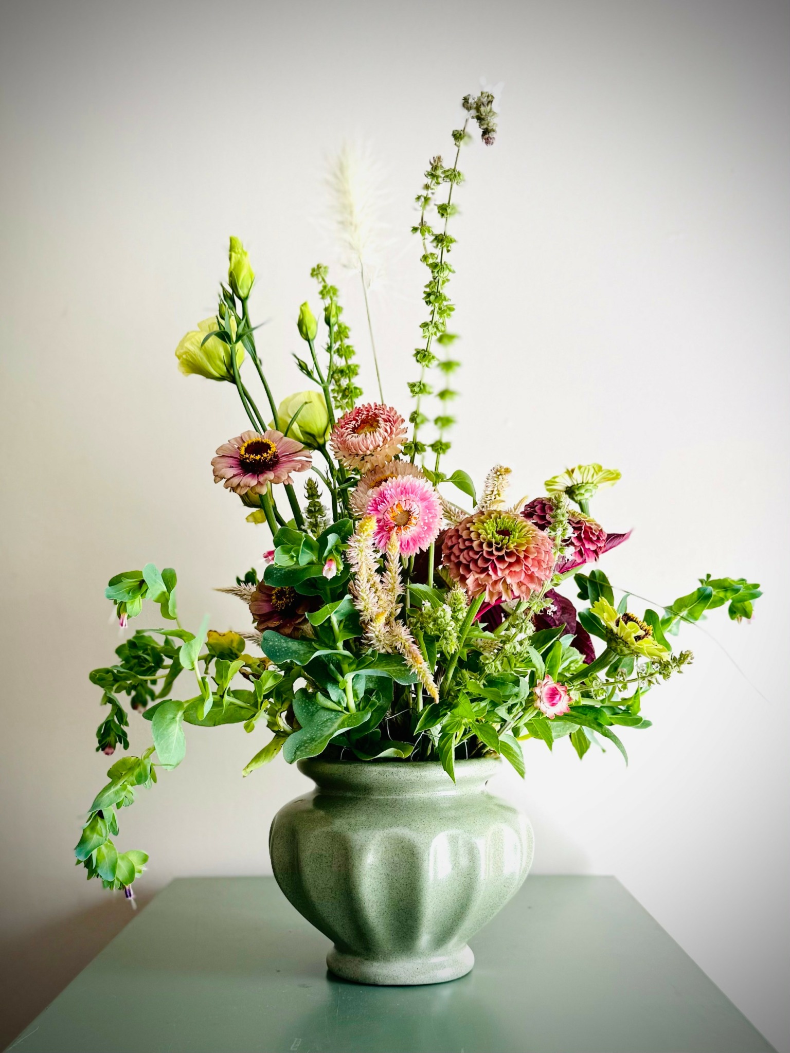 Flower arrangement with pink, green, and yellow flowers in a green ceramic vase on a white surface.
