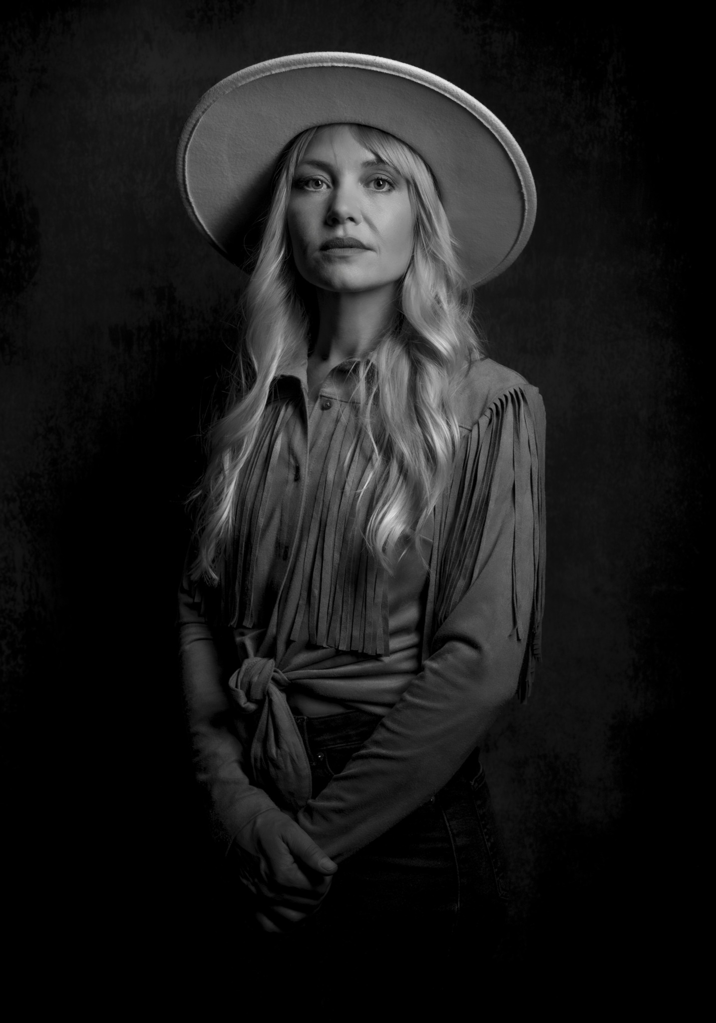 Woman with long hair wearing a wide-brimmed hat and a tied shirt, standing against a dark background.