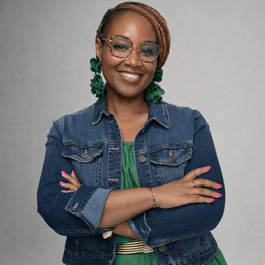 Smiling woman with glasses, braided hair, green earrings, denim jacket, and crossed arms.