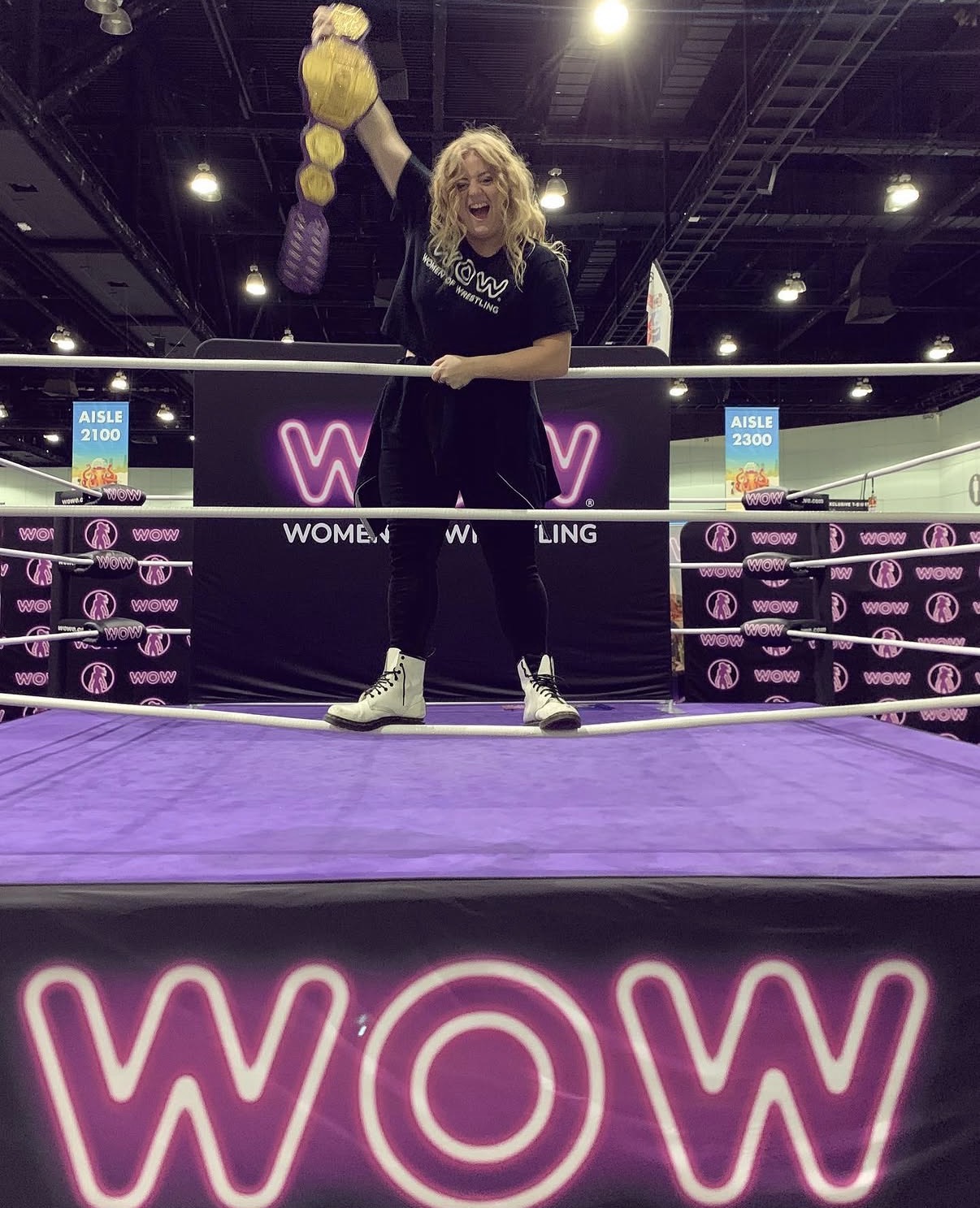 Woman with blonde hair standing on a purple stage with a large 'WOW' sign behind her, holding a pole with plush toys hanging from it.