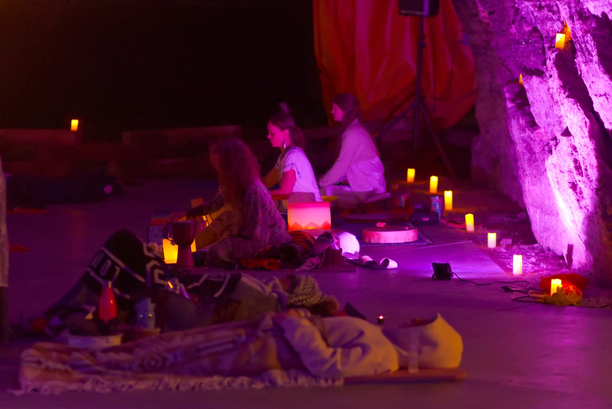 People sitting on the ground with candles and fabric, illuminated by purple and yellow lighting.
