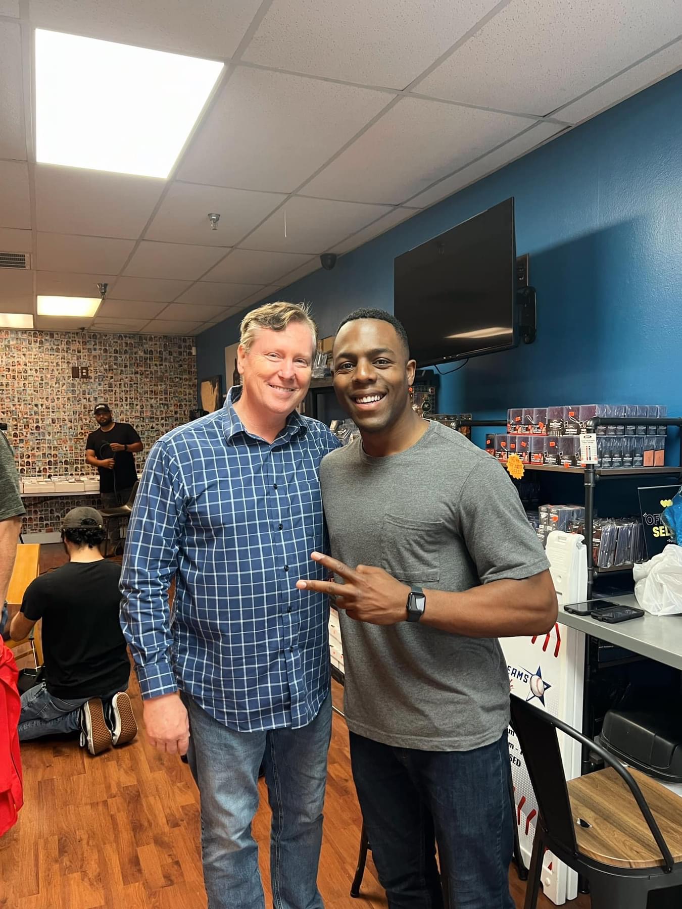 Two men standing together in a store, smiling, with shelves and a television behind them.