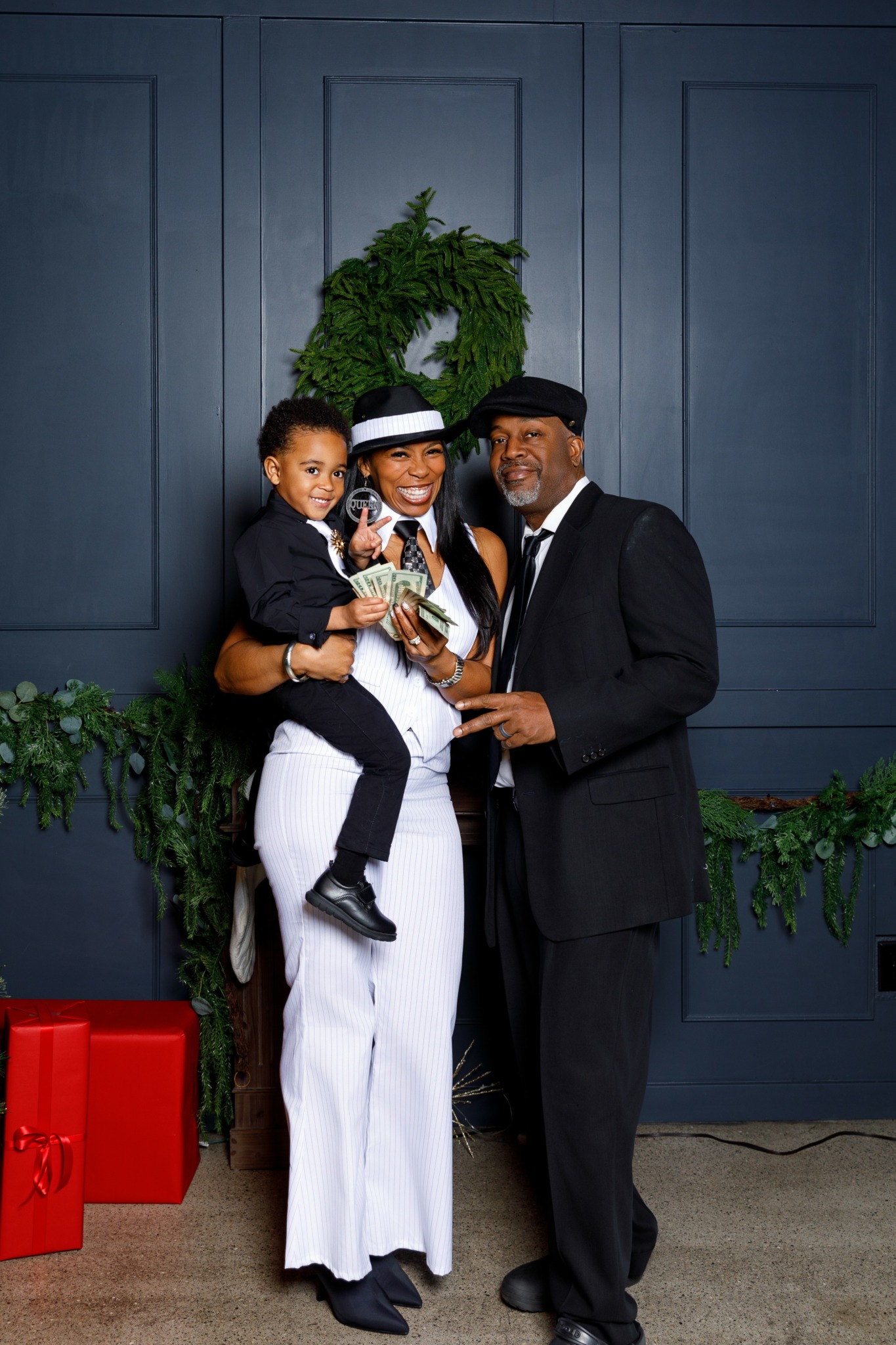 Family of three standing together, smiling, in front of a dark blue wall with greenery, holding a small dog.