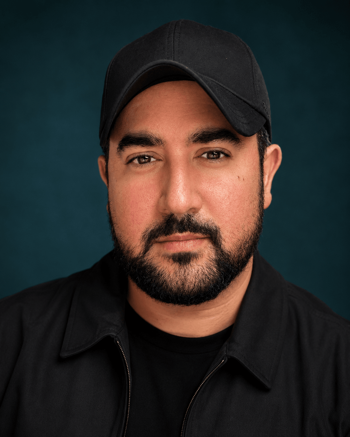 Man with a beard wearing a black cap and black shirt, facing forward, dark background.