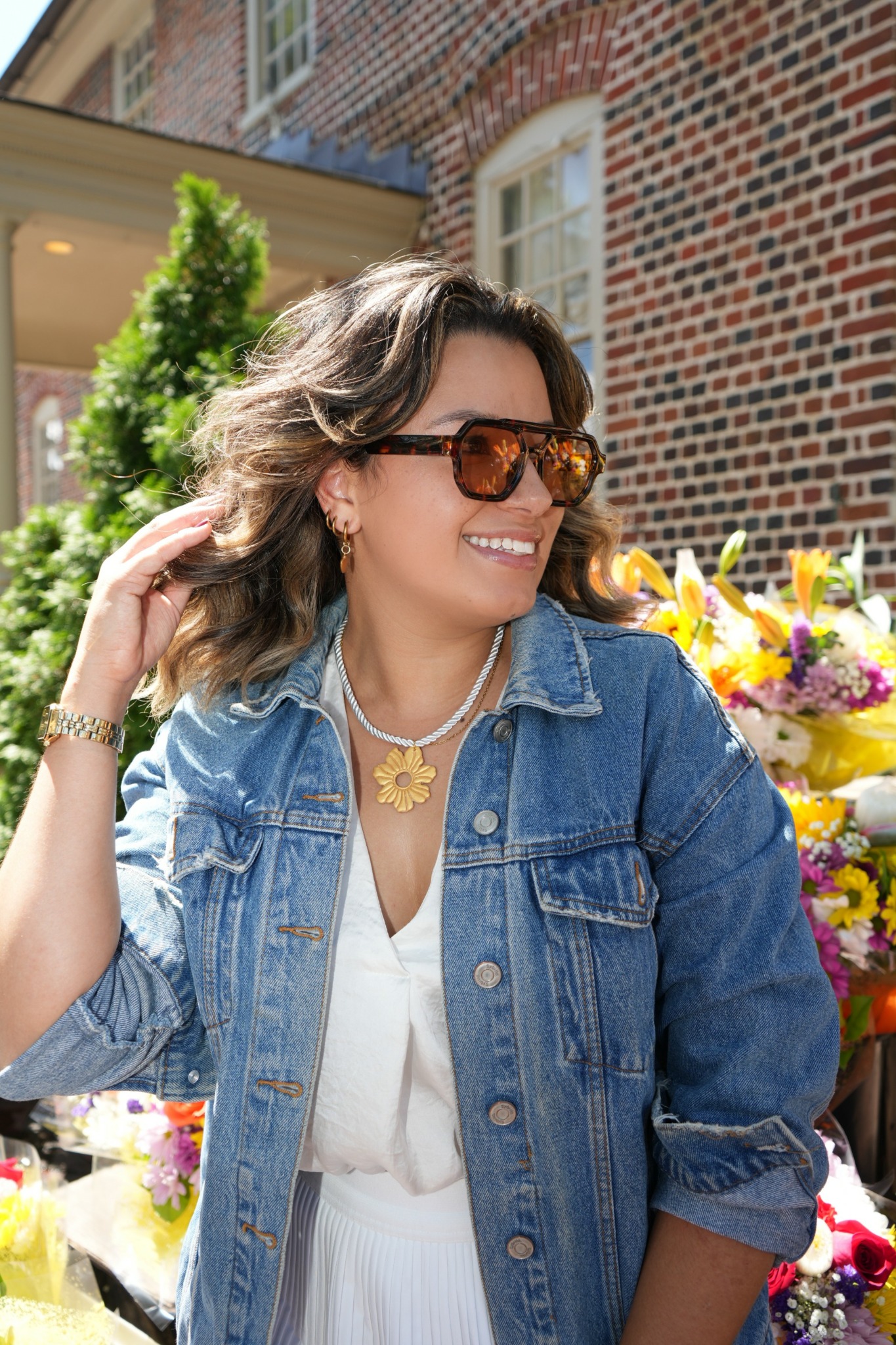 Woman with wavy hair wearing sunglasses, denim jacket, and white dress, smiling outdoors with flowers and brick building in background.