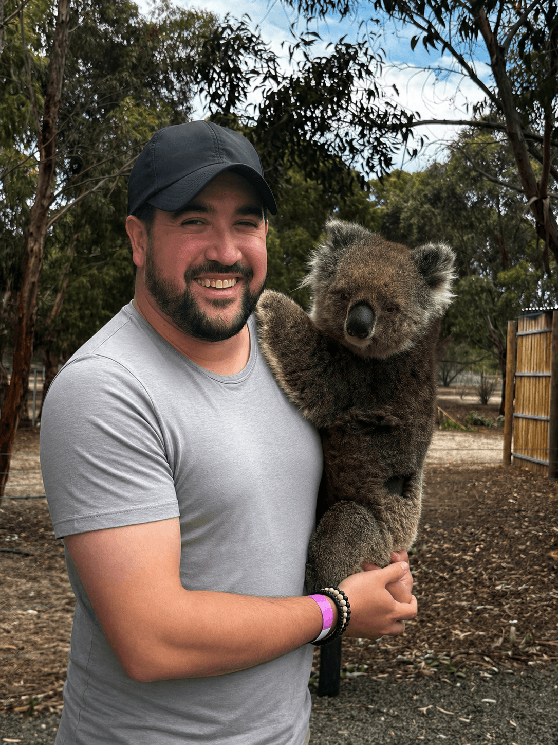 Man holding a young bear cub outdoors with trees and a wooden fence in the background.