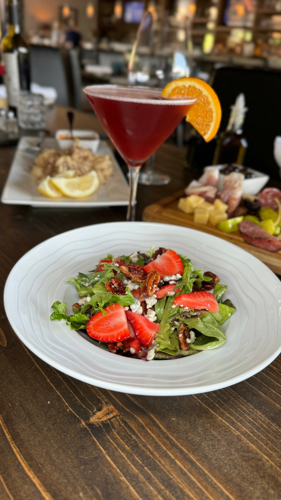 Fresh salad with strawberries and greens on a white plate, with a red cocktail garnished with an orange slice in the background.