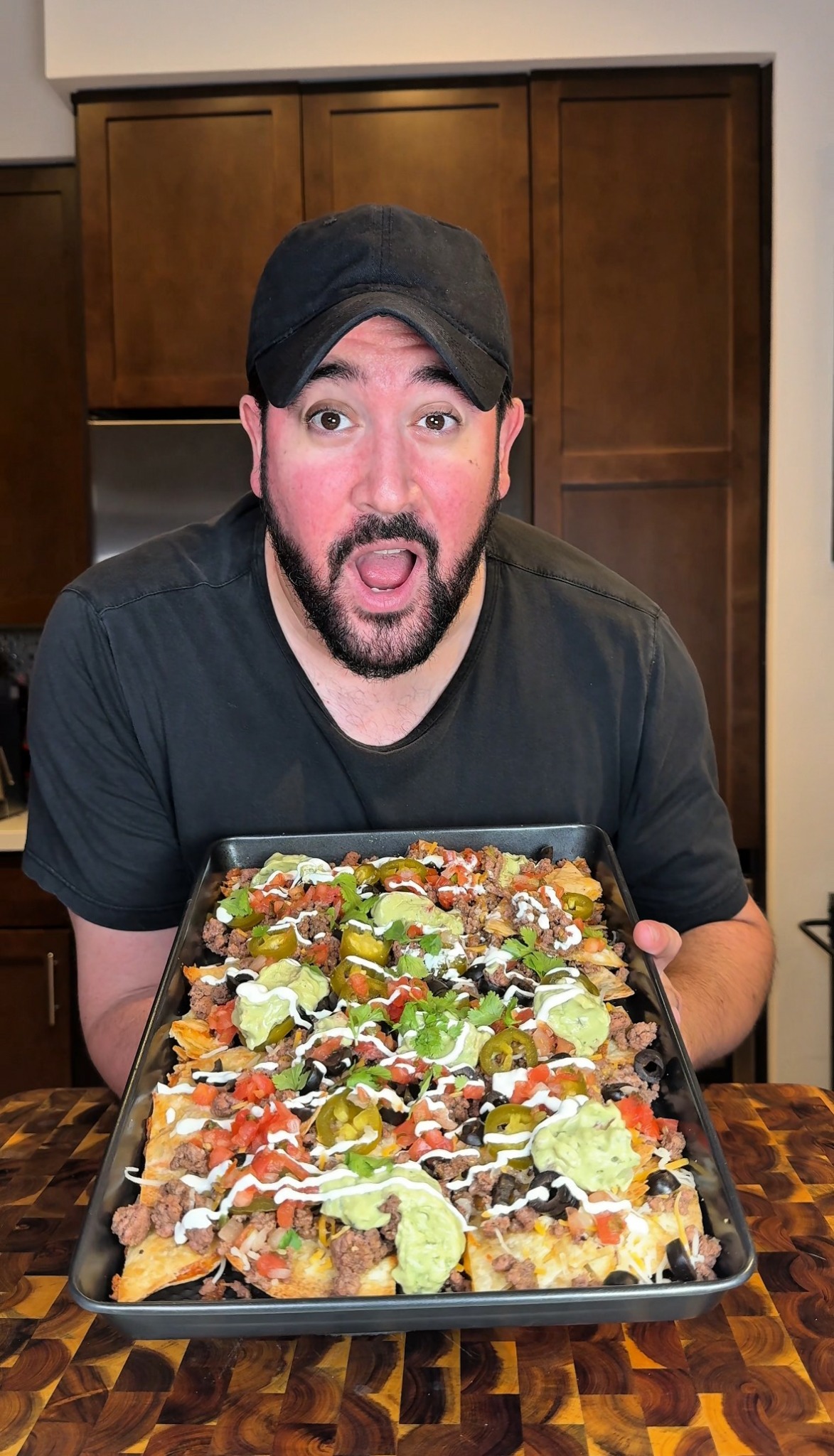 Man with a beard and black cap holding a tray of nachos topped with cheese, guacamole, and salsa, smiling.
