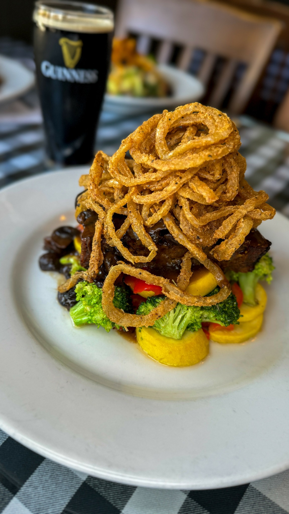 Plate with fried onion rings, vegetables, and meat, with a glass of Guinness beer in the background on a checkered tablecloth.