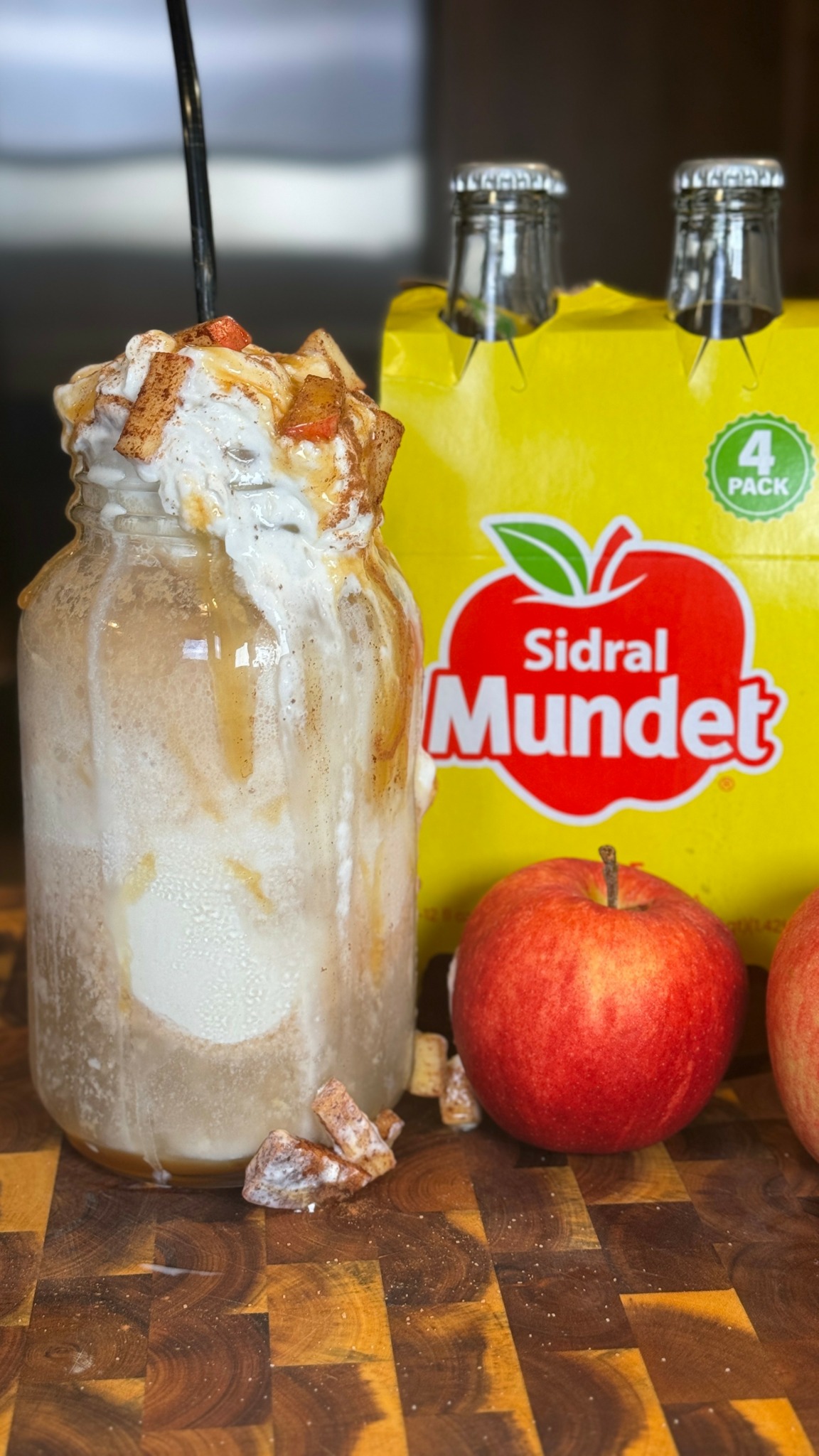 A milkshake in a glass jar topped with whipped cream, caramel, and a straw, with apple juice bottles and apples in the background.