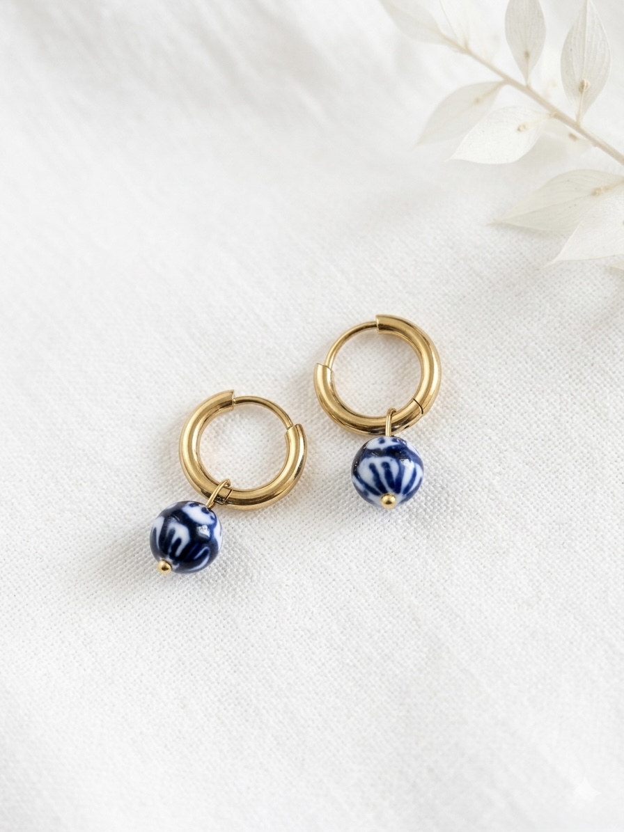 Pair of gold hoop earrings with blue and white patterned beads on a white fabric surface, with a faint leaf pattern in the background.