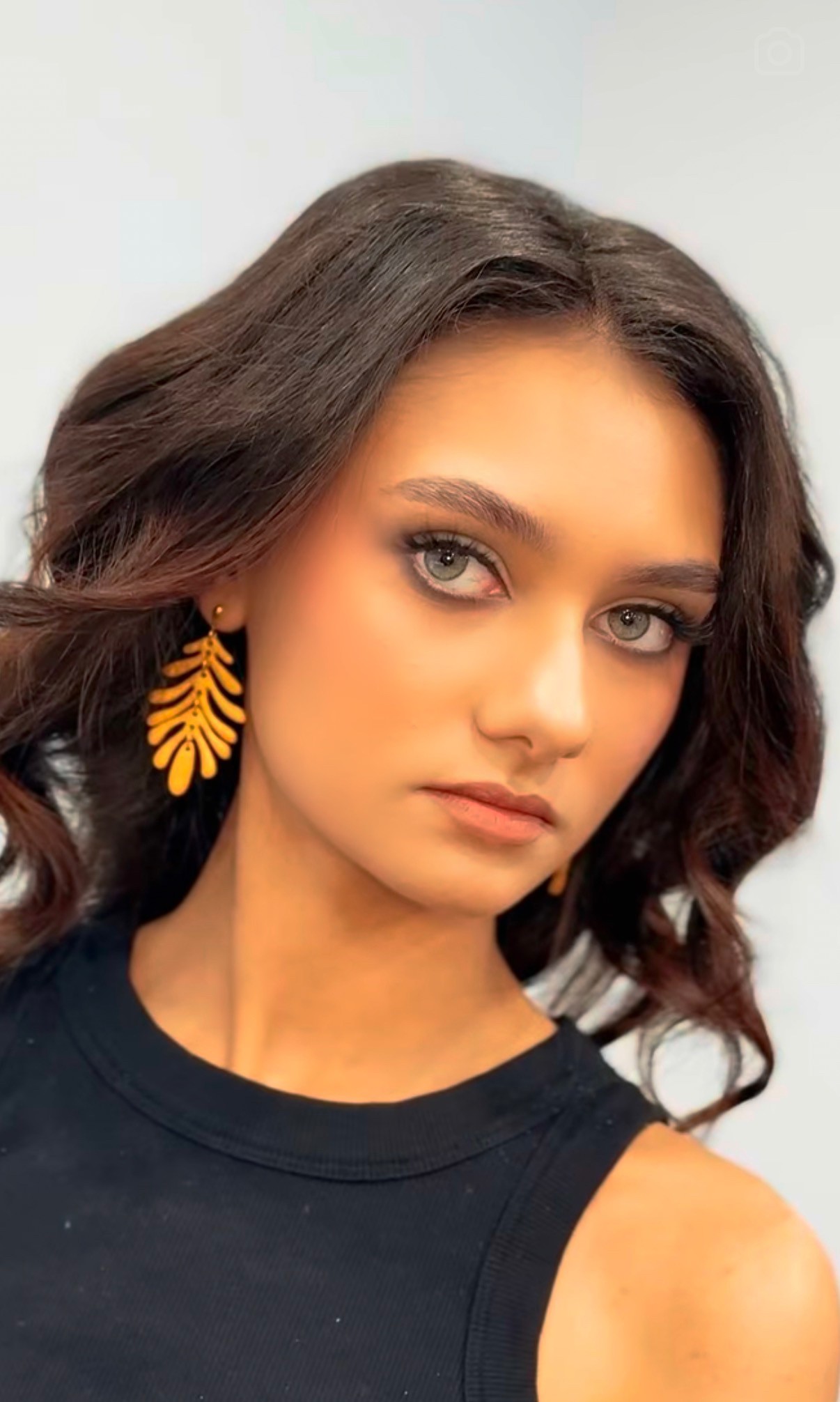 Young woman with wavy brown hair, wearing yellow leaf-shaped earrings and a black sleeveless top, looking at the camera.