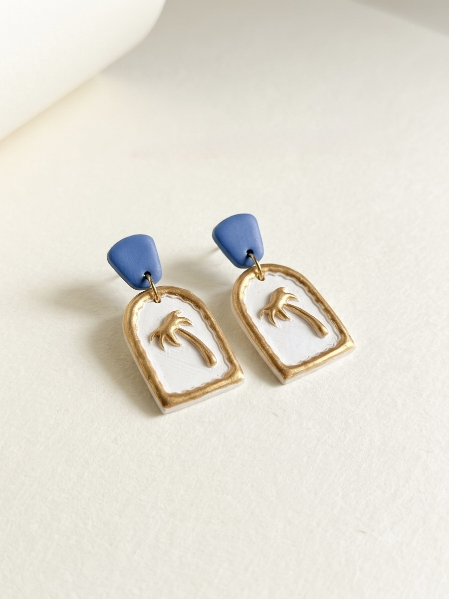 Pair of earrings with blue top parts and gold-framed rectangular pendants featuring palm tree designs, on a white background.