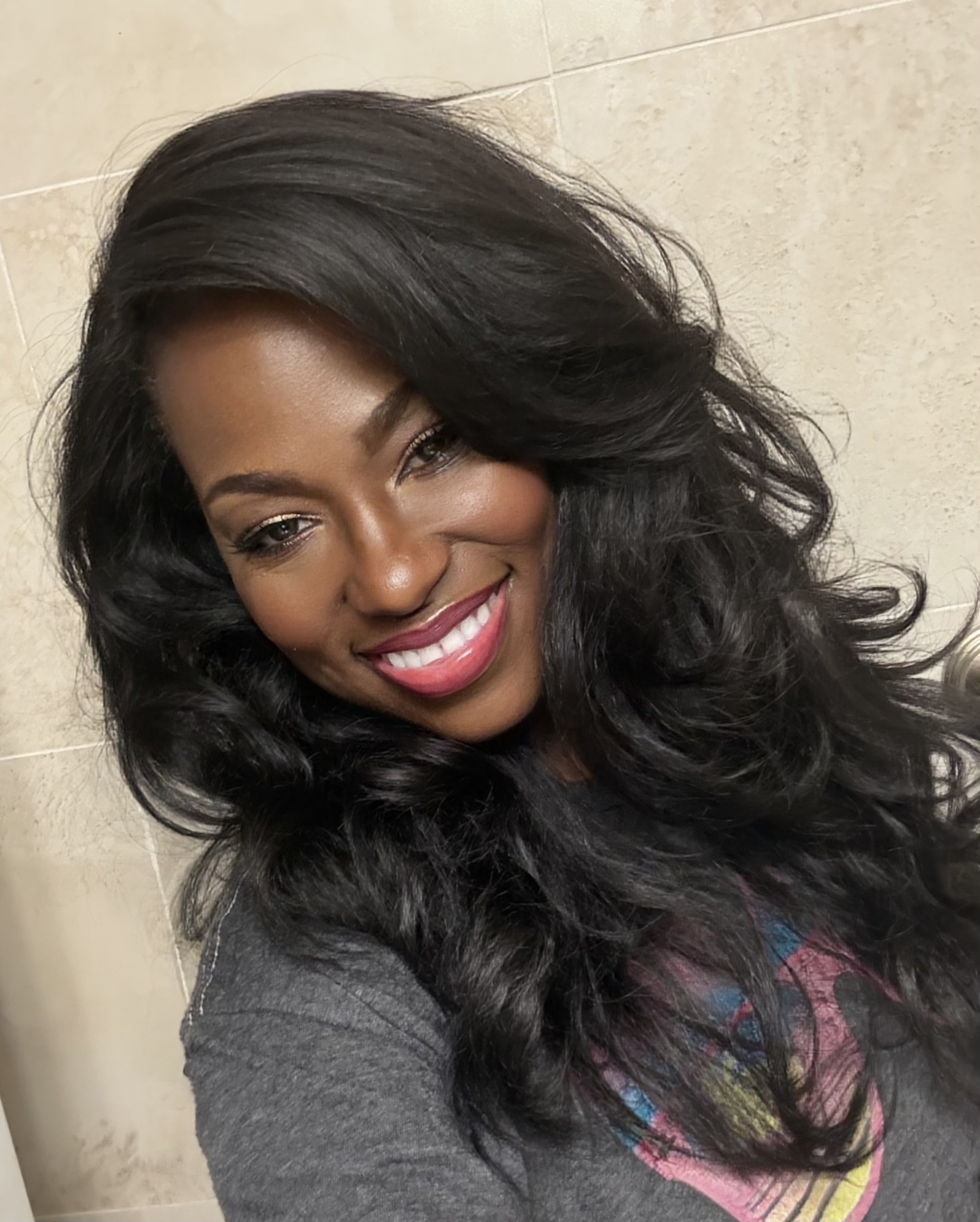 Smiling woman with long, wavy black hair, wearing a gray top, against a beige background.