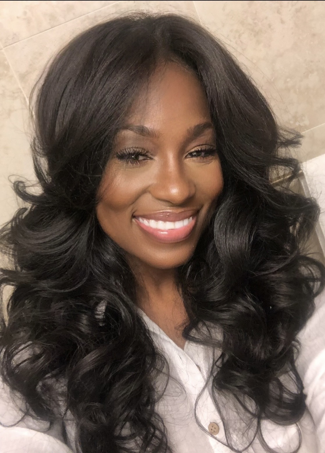 Smiling woman with long, wavy black hair and a white top, in a neutral background.