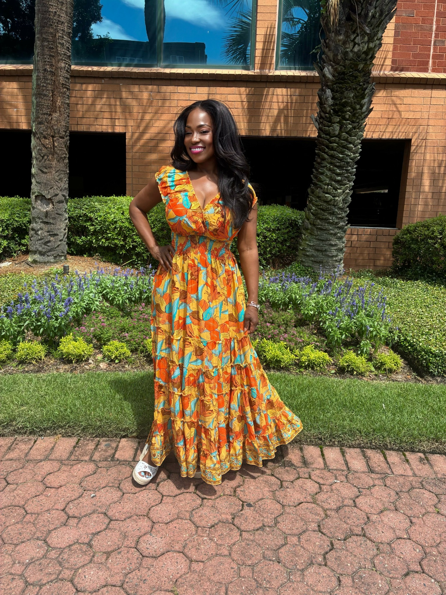 Woman in a colorful orange and yellow patterned dress standing outdoors on brick pathway, smiling, with trees and building in background.