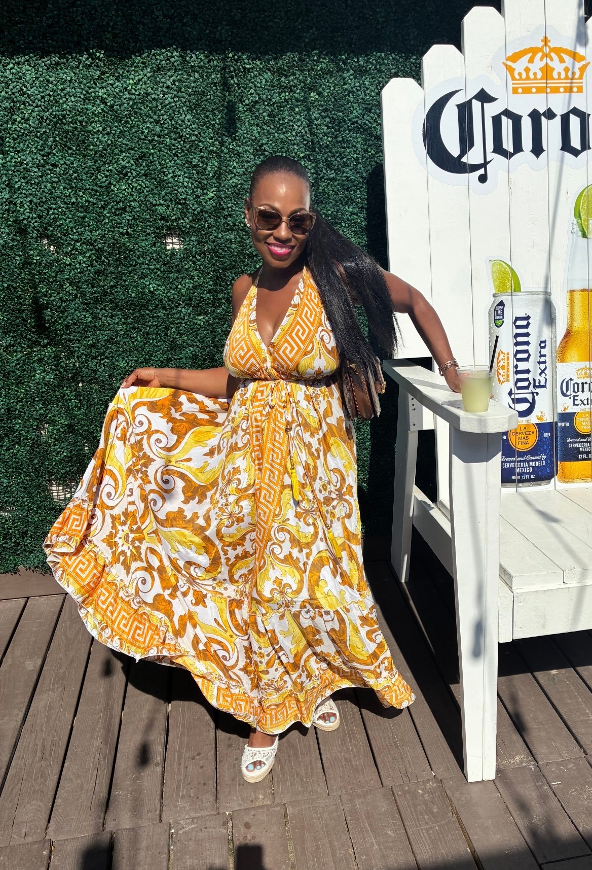 Smiling woman in a yellow and white patterned dress standing outdoors next to a white Corona-themed bar table with bottles, on a wooden deck.